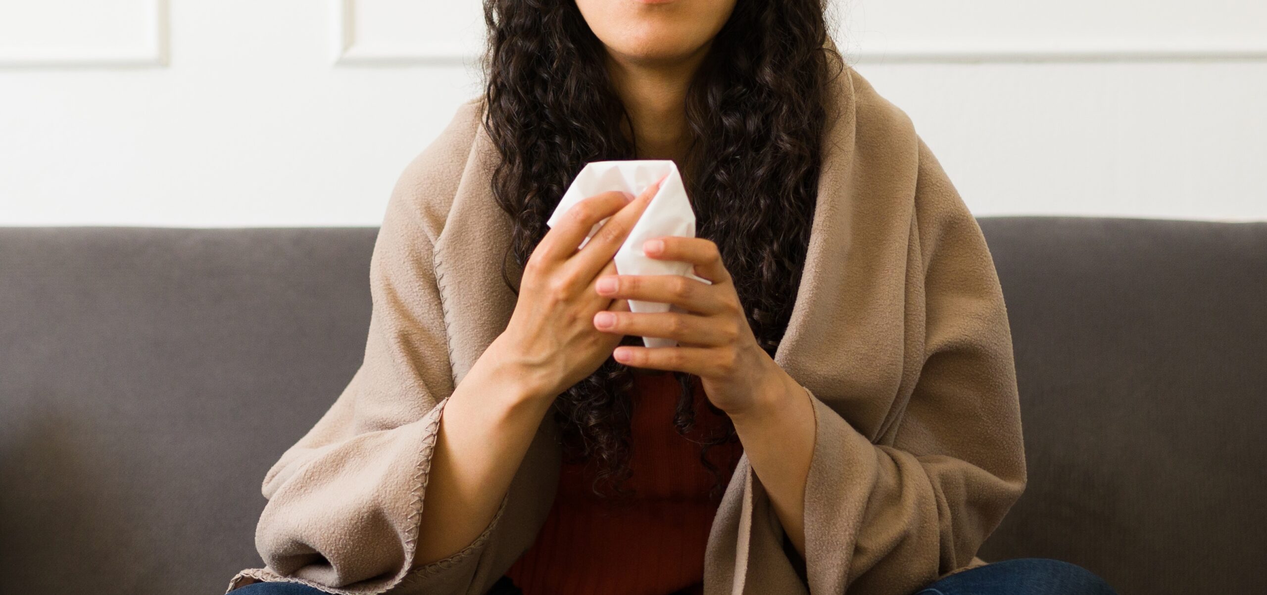 Upset sick young woman using tissues blowing her nose wrapped in a blanket while feeling sick with a bad cold or flu